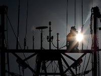 The sun rises in the distance as a painter finds himself high above New Bedford harbor using a portable high pressure air gun to remove the existing paint from the masts of a fishing boat, before giving it a fresh coat of paint.  PHOTO PETER PEREIRA