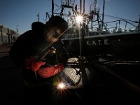 Two points of light shine through as the sun rises in the distance above, while Chris Macedo of Macedo Welding, welds a metal frame together, which will enclose the batteries of a fishing boat docked at State Pier in New Bedford.