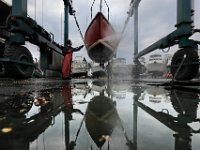 Eric Plumb is reflected in a puddle of water, as he uses a power washer to clean the underside of a sailboat recently pulled out of the water for the season at Barden's Boat Yard in Marion.