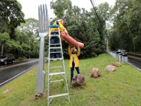 Kate Schmitt hands the hot dog to her husband Tom Schmitt who will place it atop the fork sculpture they installed at the intersection of River Road and Old Harbor Road in Westport.  They install the hot dog every year to celebrate the 4th of July.