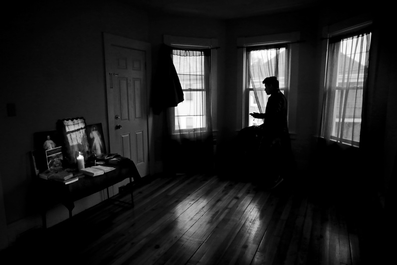 An undocumented man living in a home in New Bedford, MA looks out the window waiting for a ride to arrive.  In the corner a religious shrine can be see with a candle perpetually lit.  PHOTO PETER PEREIRA