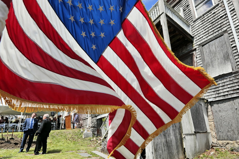 An American flag waves in the breeze as James Reed, executive director of the Veterans Transition House, speaks with Bruce Portas in front of the building seen on the right, which will be called the SFC Bruce Portas Transitional Housing Center, during the groundbreaking ceremony held in New Bedford, MA.  PHOTO PETER PEREIRA