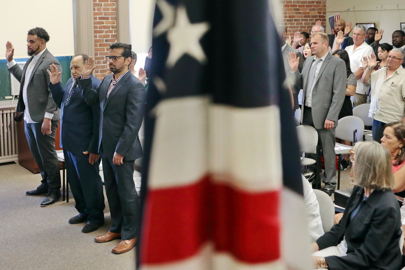 An American flag stands in the foreground as 31 new United States citizens from 12 different countries, pledge the Oath of Allegiance during the naturalization service held at the Immigrants Assistance Center on Crapo Street in New Bedford, MA.   PHOTO PETER PEREIRA