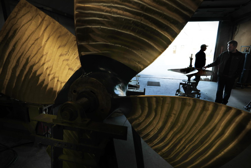 Mihkel Valm and Andrius Limantas move a fishing boat propeller which needs to be repaired onto a jig, while in the foreground sits a monster propeller from another dragger that they recently repaired at the Fleet Propeller shop in Fairhaven, MA.