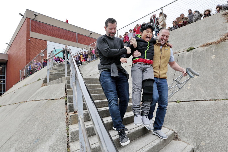 Hayden McFadden teachers, Michael Roderick, left, and physical education teacher Brian Coon, give fifth grader, Daniel Bejarano Alvarado, who injured his knee outside of school,  a hand going down the stairs, so that he could be closer to his classmates during the egg-drop in front of their school in New Bedford, MA.