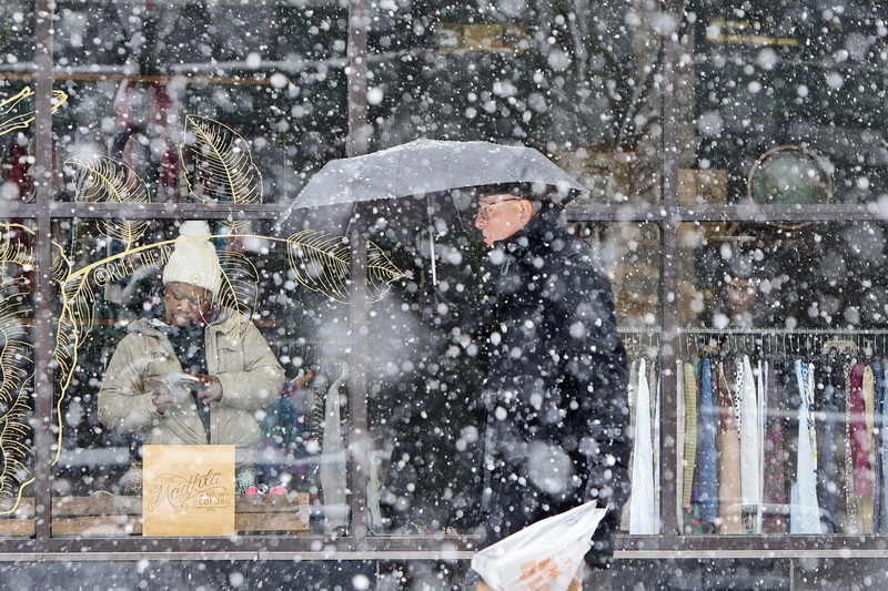 A man with umbrella in hand walks past The Madlita in downtown New Bedford, MA as snow continues to fall, while inside a woman takes a look at a pair of shoes on sale.  PHOTO PETER PEREIRA