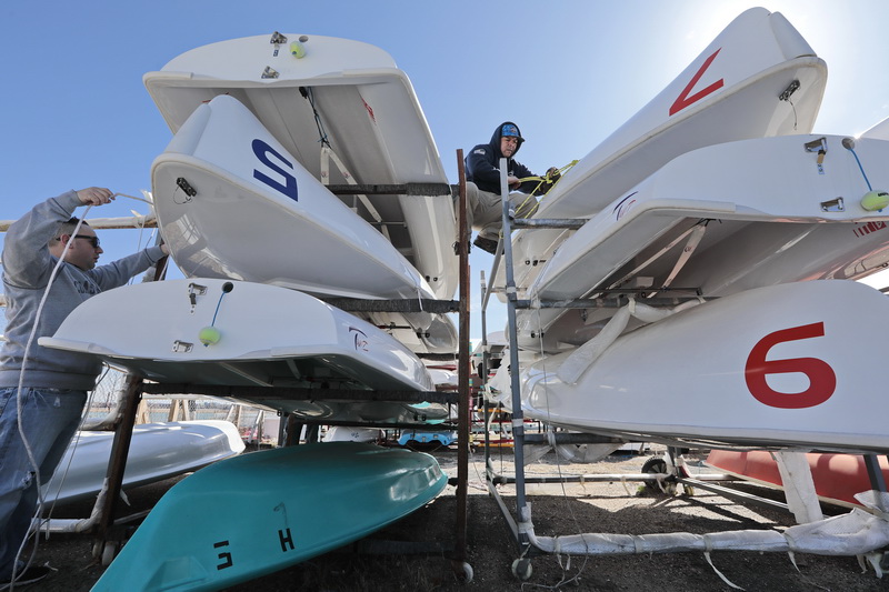 Coast Guardsmen SCPO Manny Gonzales (Woods Hole, top) and SCPO Ryan Vess, left, remove some of the collegiate 420 boats from their winter storage racks, in preparation for next week when Stang and UMass Dartmouth start their sailing season. Coast Guardsmen from various stations across the region, volunteer their time at the Community Boating Center to help the New Bedford non-profit prepare for the upcoming sailing season.   PHOTO PETER PEREIRA