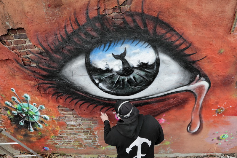 Graffiti artist Jeff St.Pierre works on a mural featuring a crying eye with the icons of the whaling city reflected in its pupil, surrounded by coronavirus spores.  He and fellow graffiti artist, Ryan McFee, are painting the piece on a building adjacent to the Custom House Square in New Bedford with the hope of eventually replacing the spores with flowers are the virus is defeated.   PHOTO PETER PEREIRA