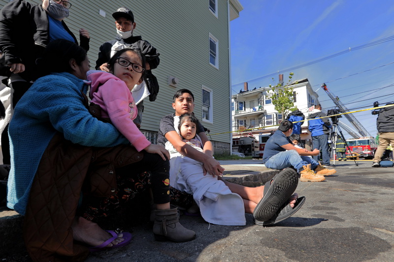 Displaced families wrap themselves with blankets given to them by New Bedford EMS as they sit on the sidewalk. In the background New Bedford firefighters can be seen battling a three-alarm fire at 371 N. Front Street in the north end of New Bedford. Buildings on both N. Front Street and Tallman Streets were affected by the blaze. PHOTO PETER PEREIRA