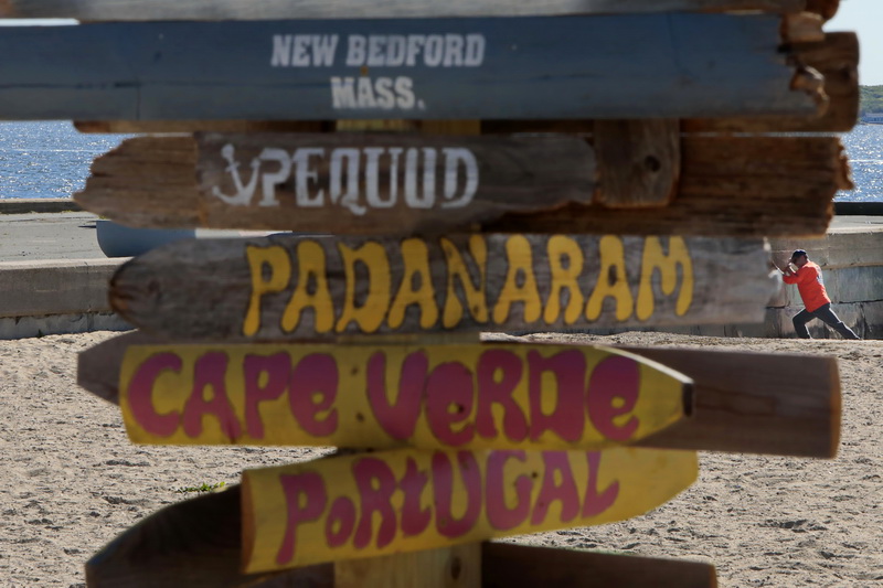 From this perspective, a man doing stretches against the pier wall, looks like he thinks the Padanaram sign needs to be pointed in a new direction, on a set of signs pointing to different parts of the city and the world installed on East Beach in New Bedford, MA.  