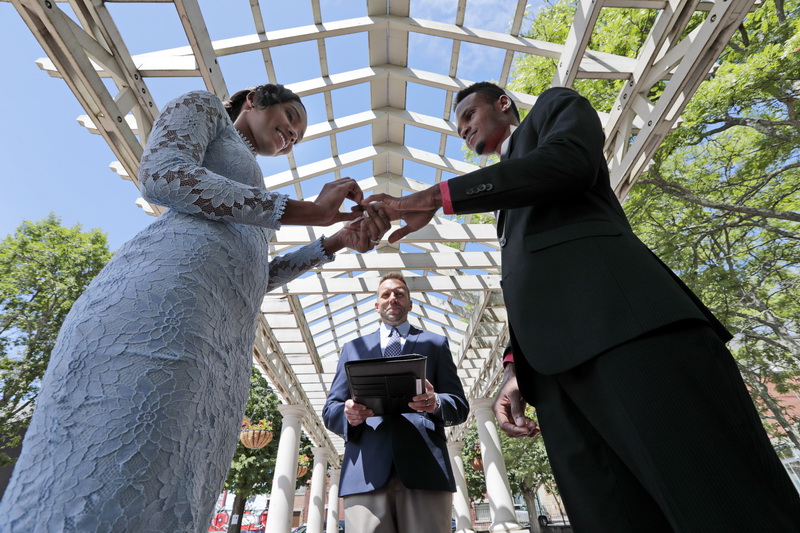 New Bedford City Clerk, Dennis Farias looks on, as Penelope Bazile places a wedding ring on Gligorov Bazile's finger during their wedding held at Custom House Square in downtown New Bedford, MA.  With weddings not allowed inside City Hall since the pandemic shutdown in March, City Clerk Farias has started to hold the ceremonies once again, but this time outside.  PHOTO PETER PEREIRA