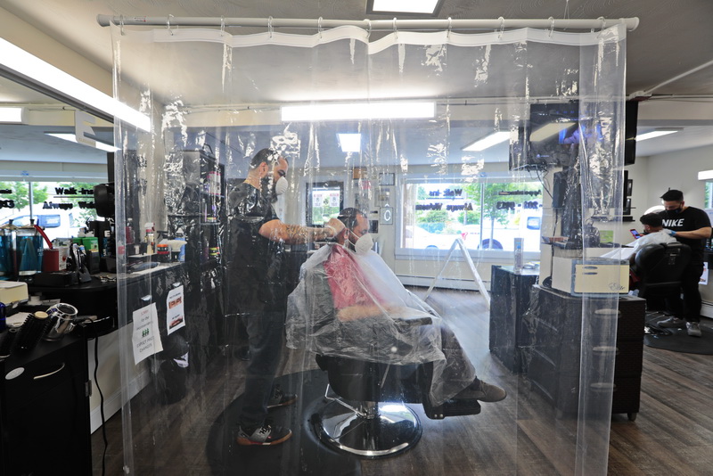 Sandro Quiles gives Gus Bedoya a haircut as seen behind a clear screen erected between the stations for protection, at the Upper Kutz barber shop on Rockdale Avenue in New Bedford, MA.  PHOTO PETER PEREIRA