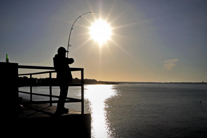 The sun rises in the distance as Antonio Carreiro casts from the top of the hurricane barrier in New Bedford, MA.  PHOTO PETER PEREIRA