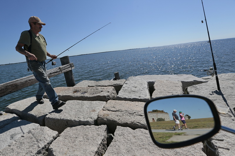 Stanley Bisaillon walks over to put some fresh bait on the hook while from a pier at Fort Taber Park in the south end of New Bedford, MA while in the mirror of his motorized bicycle, a couple can be seen wallking the paths behind Fort Rodman. 