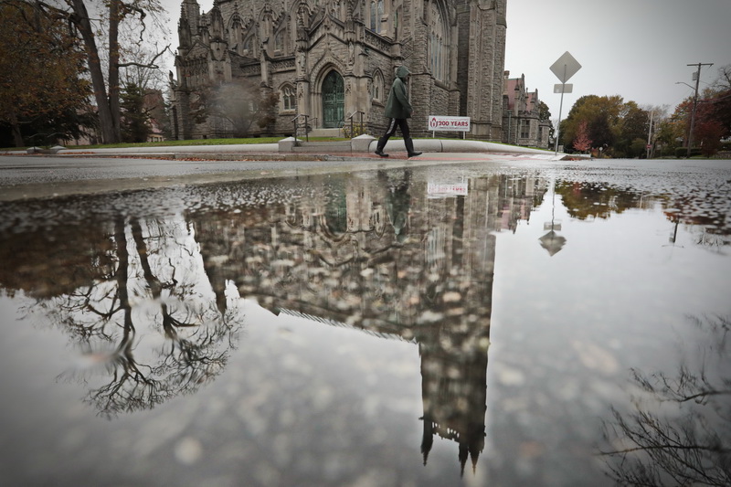 The steeple of the Unitarian Church in Fairhaven, MA is reflected in a puddle, as a man makes his way up Green Street for a rainy walk.   PHOTO PETER PEREIRA