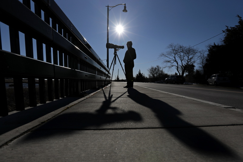 With the sun rising in the distance the shadow of wildlife photographer David Jeffrey and his telephoto lens are displayed on the sidewalk as he waits for the perfect moment to photograph hooded merganser ducks he spotted in the distance in Padanaram Harbor in Dartmouth.  Mr. Jeffrey is the author of Birds: Intimate Images a book featuring many birds from our area and in Florida.  The book is available on Amazon and Barnes & Noble. PHOTO PETER PEREIRA 