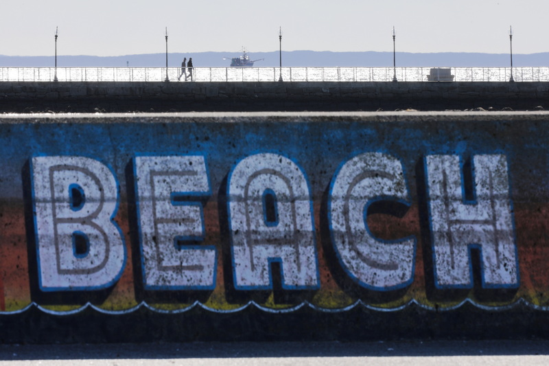 Two men go for a walk atop the pier at Fort Taber Park as a fishing boat heads out to sea in the south end of New Bedford.  In the foreground the East Beach sign painted on the side of the concrete barrier between the street and the sand. PHOTO PETER PEREIRA