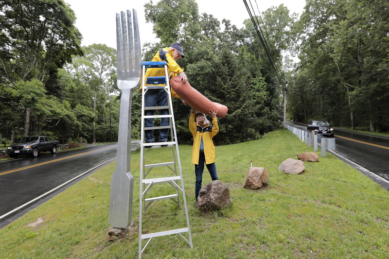 Kate Schmitt hands the hot dog to her husband Tom Schmitt who will place it atop the fork sculpture they installed at the intersection of River Road and Old Harbor Road in Westport.  They install the hot dog every year to celebrate the 4th of July. PHOTO PETER PEREIRA