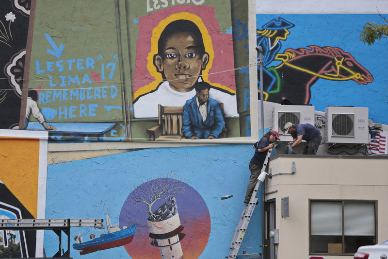 Roofers have become part of the Postcards from New Bedford murals painted on the side of a building downtown, as they replace the roof for the Bank of America parking attendant.