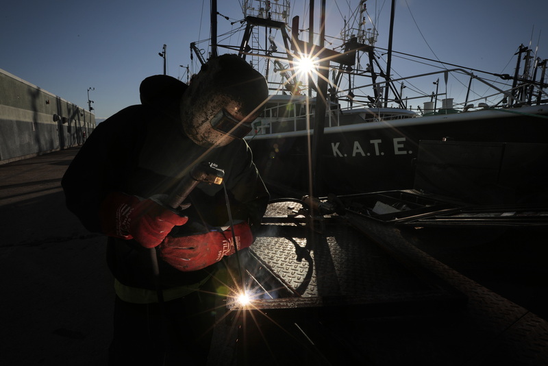 Two points of light shine through as the sun rises in the distance above, while Chris Macedo of Macedo Welding, welds a metal frame together, which will enclose the batteries of a fishing boat docked at State Pier in New Bedford, MA.  PHOTO PETER PEREIRA