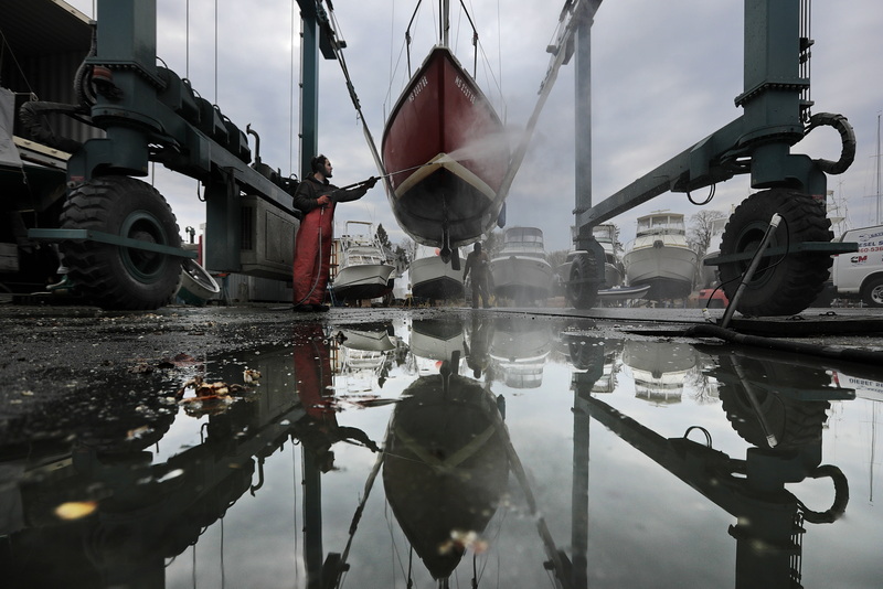 Eric Plumb is reflected in a puddle of water, as he uses a power washer to clean the underside of a sailboat recently pulled out of the water for the season at Barden's Boat Yard in Marion, MA. PHOTO PETER PEREIRA