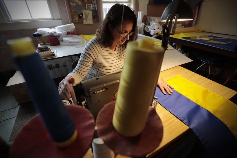 Dianne Veiga of Brewer Banner Designs is seen between blue and yellow spools of thread, as she sews a Ukrainian flag for a customer who was looking to show support for Ukraine but was unable to find any.  All proceeds from the sale of these flags will be donated to Medecins sans Frontiers by the custom New Bedford, MA flag maker. PHOTO PETER PEREIRA