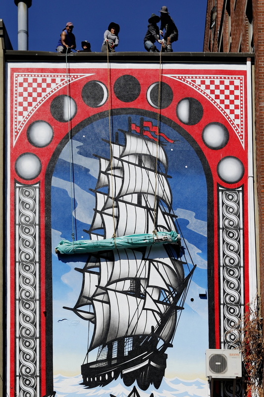 Roofers hoist a roll of rubber roofing past the whaling ship mural painted on the side of a building at Wings Court in New Bedford, MA. PHOTO PETER PEREIRA