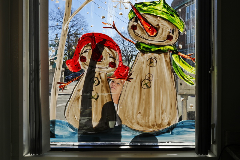 Tuesday Desrochers is seen between the two snowmen she is painting on the window of the Goulart Square Bakery on Rivet Street in New Bedford, MA as the shadow of her arm drawing the hat of the smaller snowman, is cast on the window.