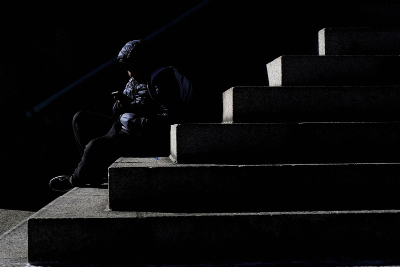 A sliver of light illuminates a man looking at his smartphone and the steps he is sitting on in front of the New Bedford public library on Pleasant Street. PHOTO PETER PEREIRA