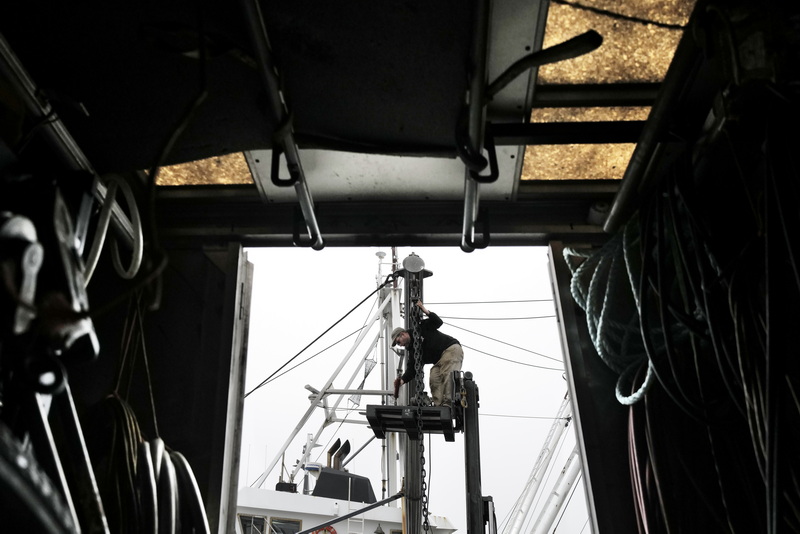 Scott Cardin is elevated on a lift while he works on repairing the outrigger chain of the fishing boat Concordia, which is moored at Union Wharf in Fairhaven, MA as observed from the tool van. PHOTO PETER PEREIRA