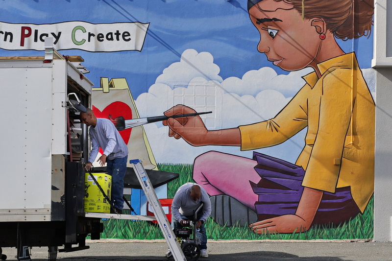 The mural depicting a young girl on the wall of Little Peoples College on Dartmouth Street in New Bedford, MA appears to be painting Melvin Santiago as he and Angel Santos take their equipment out of the back of a truck prior to power washing the building. PHOTO PETER PEREIRA