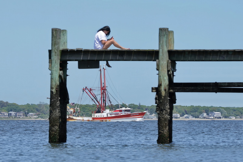 A woman enjoys the weather atop a pier in New Bedford, MA as the fishing boat Instigator heads out to sea in the distance. PHOTO PETER PEREIRA