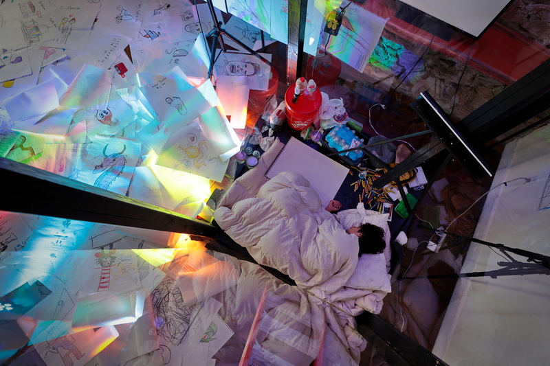 Mason Terra, a local artist, takes a quick nap during the twenty-four hour period in which he confined himself in a glass box at the Co+ Creative Center in downtown New Bedford, MA equipped only with drawing materials and water. Throughout this time, viewers could ask Mason to create drawings through a livestream on YouTube.  PHOTO PETER PEREIRA