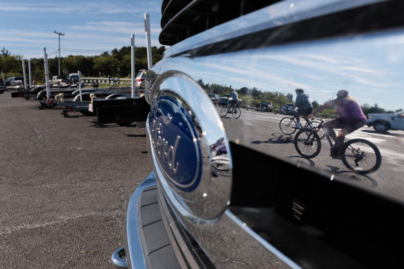 Three cyclists enjoy the weather and the view as they are reflected on the grill of one of the pickup trucks hauling a boat trailer parked at the Westport, MA River boat ramp. PHOTO PETER PEREIRA