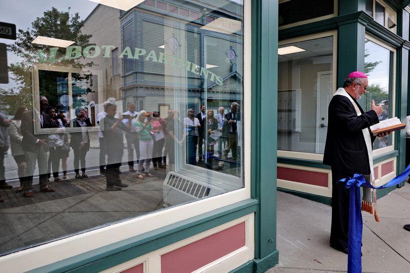 Guests are reflected on the storefront windows of the Talbot Apartments on Acushnet Avenue in New Bedford, MA as Bishop Edgar M. da Cunha offers prayers to the future apartment complex. PHOTO PETER PEREIRA