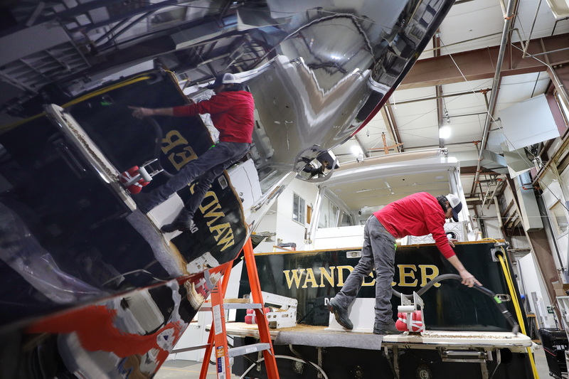 Eric Parker�s reflection appears on the hull of a nearby boat while he vacuums up the wood chips produced from routing the wood trim of the Wanderer prior to applying varnishing at Concordia in Dartmouth, MA. PHOTO PETER PEREIRA