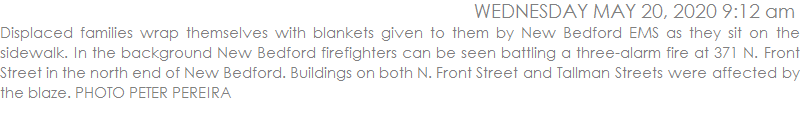 Displaced families wrap themselves with blankets given to them by New Bedford EMS as they sit on the sidewalk. In the background New Bedford firefighters can be seen battling a three-alarm fire at 371 N. Front Street in the north end of New Bedford. Buildings on both N. Front Street and Tallman Streets were affected by the blaze. PHOTO PETER PEREIRA