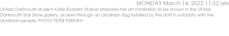 UMass Dartmouth student Katie Rodden Walker prepares her art installation to be shown in the UMass Dartmouth Star Store gallery, as seen through an Ukrainian flag installed by the staff in solidarity with the Ukrainian people.