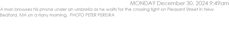 A man browses his phone under an umbrella as he waits for the crossing light on Pleasant Street in New Bedford, MA on a riany morning.  PHOTO PETER PEREIRA