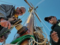 Joe Mello, left, and Jason Costa, right, of Triad Boatworks remove the rigging from the mast of a sailboat docked in Mattapoisett, as Silas Costa looks down from above as he sets up a strap to remove the mast.