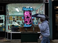 Danny Baptista, owner, gives Alex Buchanan a haircut inside of the Stepping Razor barber shop on Purchase Street in New Bedford, as a man takes shelter from the driving rain with an umbrella as he walks past outside.