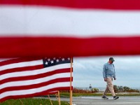 A man walks past a series of American flags placed on the edge of Fort Street at Fort Phoenix in Fairhaven.  In the background the Butler Flats lighthouse can be seen.