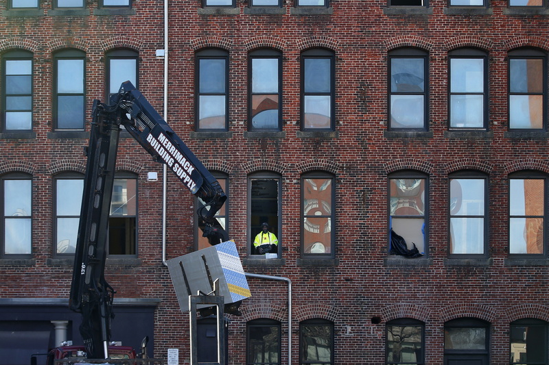 A Merrimack Building Supply worker can be seen guiding a lift filled with drywall sheets using a remote control, from the window of a building undergoing renovations in downtown New Bedford. 