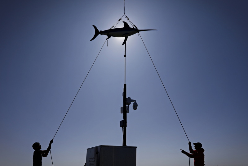 Silas Costa, left, and Jason Costa, of Triad Boatworks, use lines to align the iconic swordfish weathervane as it is lowered onto the mast on the wharf in Mattapoisett, MA. PHOTO PETER PEREIRA