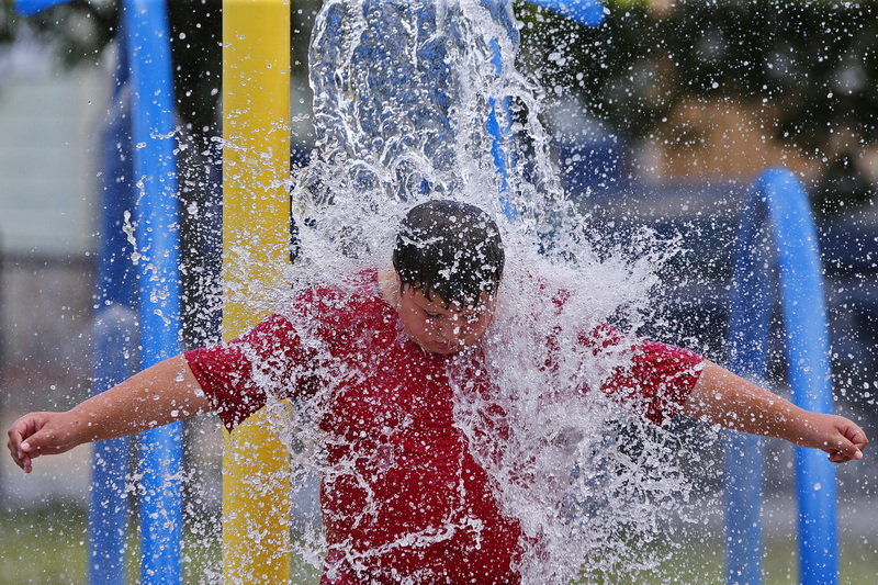 Graiden Therrien, 11, takes shelter from the sweltering heat by waiting for the automated water bucket to overturn onto his head at Harrington Park in New Bedford, MA. PHOTO PETER PEREIRA