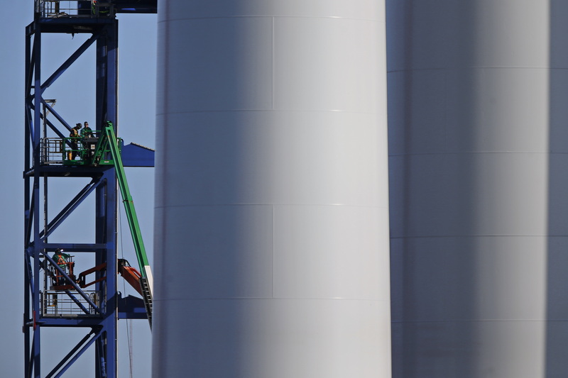 Workmen constructing a steel structure are dwarfed by two wind turbine towers being assembled at the New Bedford Marine Commerce Terminal in New Bedford. The towers will be used by Vineyard Wind for their offshore wind farm being constructed off Martha's Vineyard. PHOTO PETER PEREIRA