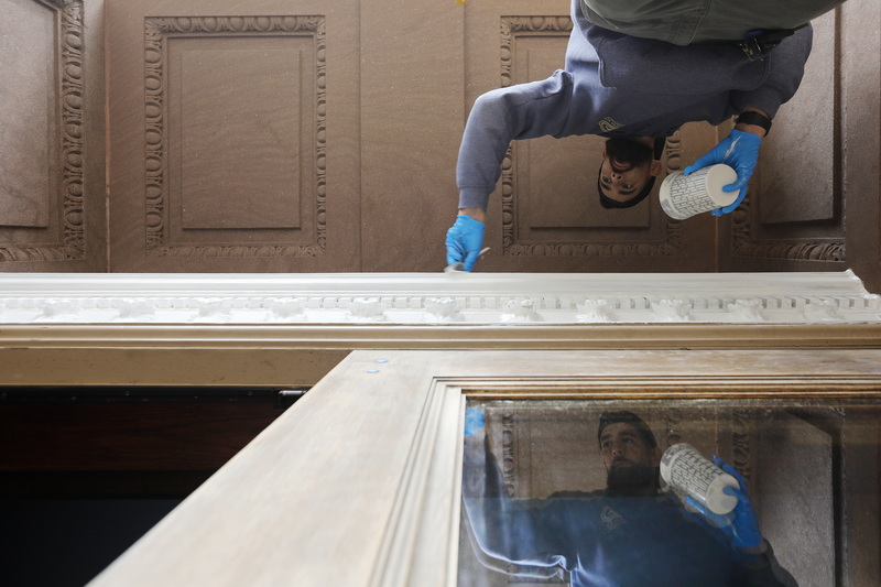 Nick Dunn is reflected on the glass of the front door of New Bedford City Hall as he paints the decorated transom above before applying stain to the door. PHOTO PETER PEREIRA