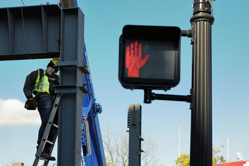 The crosswalk sign offers a hand to a worker installing a steel beam for the future residential complex being built at the intersection of Union Street and South Second Street in downtown New Bedford, MA. PHOTO PETER PEREIRA