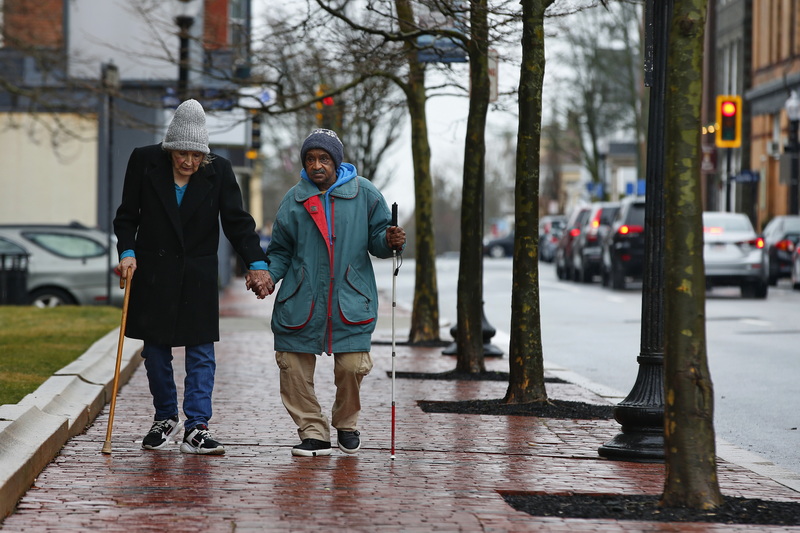 A woman gives a man with a vision impairment a hand walking down the sidewalk on N Sixth Street in downtown New Bedford, MA. PHOTO PETER PEREIRA