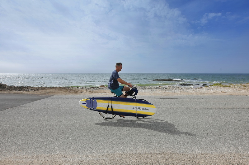 A man carries his surfboard on his bicycle as he rides down East Beach Road in Westport, MA with high surf expected across the region. PHOTO PETER PEREIRA